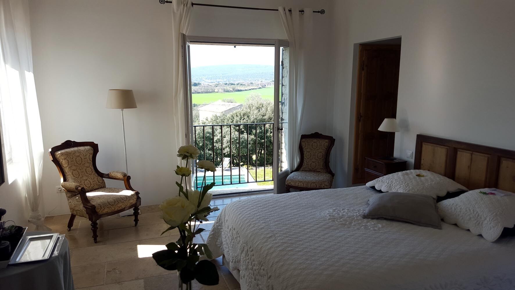 A bedroom with a large bed, two chairs, a balcony with a view, and a window with curtains.