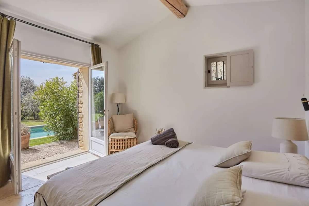A bedroom with a bed, a chair, a lamp, and a view of a garden and pool through a glass door.