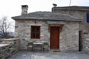 A stone building with a wooden door and window, two chairs outside, and a stone roof. - Nikolas Guesthouse (Zagori)
