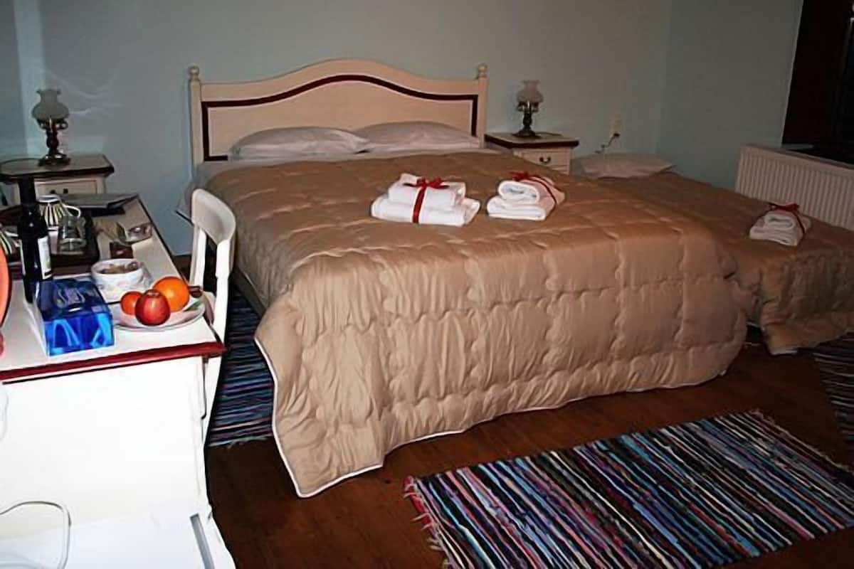 Quarto de hotel com uma cama grande, uma mesinha de cabeceira com abajur, uma pequena mesa com uma cesta de frutas e uma garrafa, e um tapete listrado e colorido.