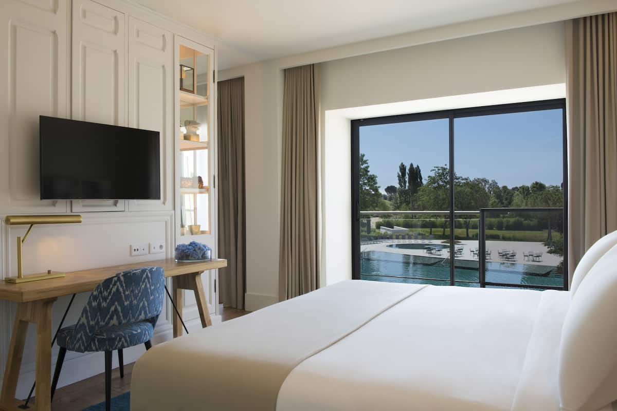 Une chambre d’hôtel avec un grand lit, un bureau avec une télévision, une chaise et une vue sur une piscine.