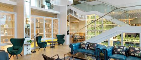 A spacious interior with a high ceiling, a central seating area with sofas and chairs, a staircase, and glass-fronted display cases.