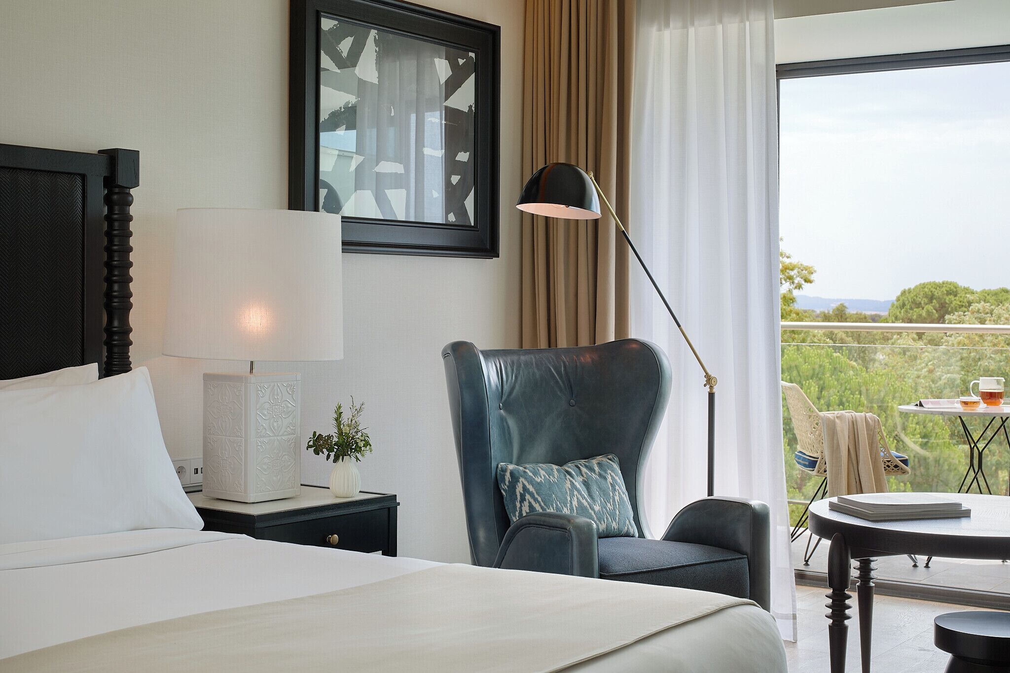 Habitación de hotel con cama, una silla, una mesa, lámpara y vista a zonas verdes.