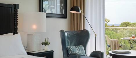 Habitación de hotel con cama, una silla, una mesa, lámpara y vista a zonas verdes.