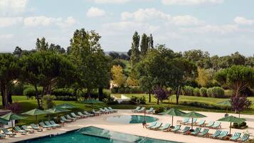 Zona de piscina con sillas de descanso y sombrillas, rodeada de árboles y vista a un campo de golf.