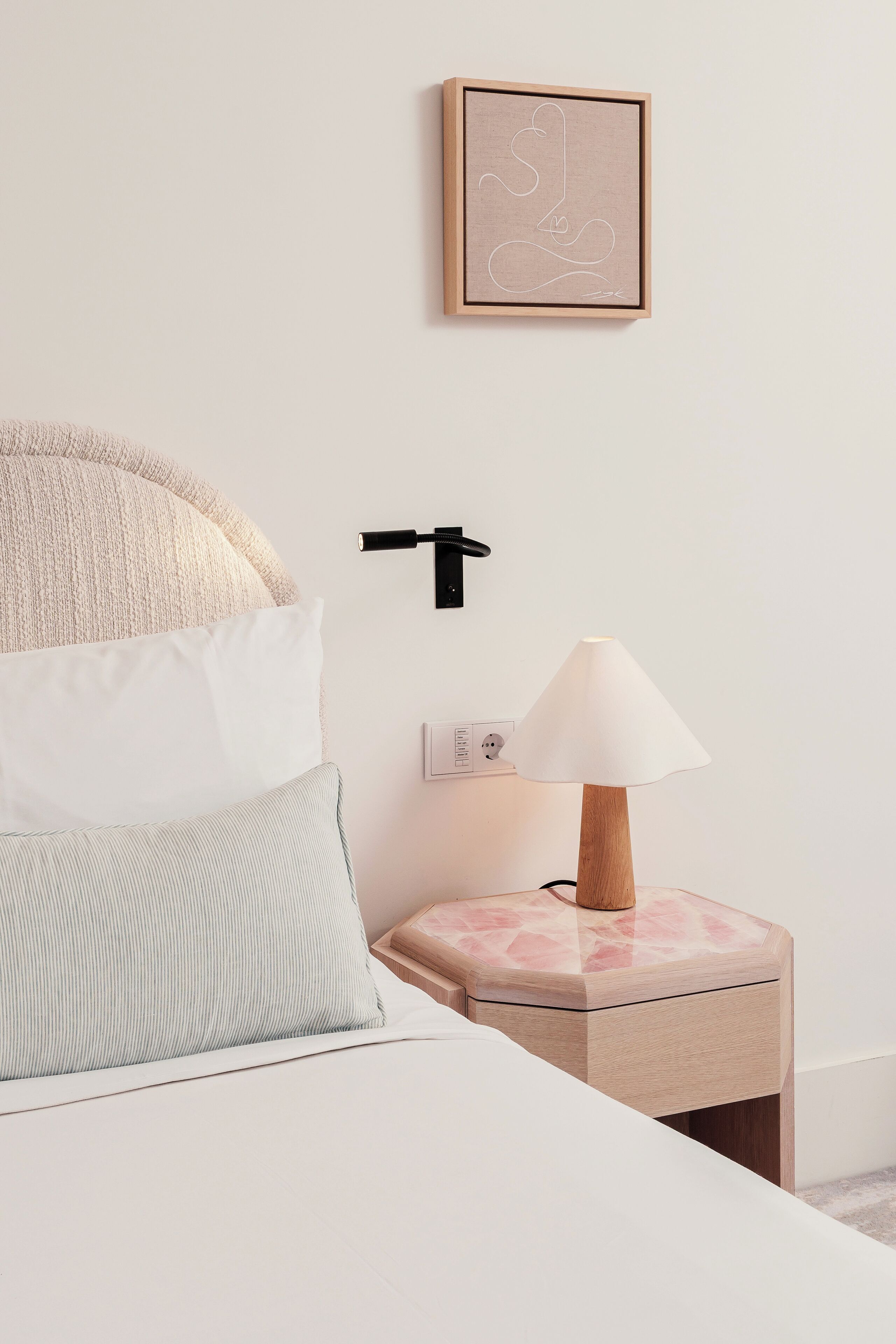 Un dormitorio ordenado con cama, almohadas, una mesita de noche con lámpara, un interruptor en la pared y una obra de arte enmarcada.