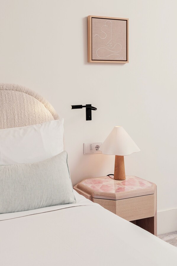 A neatly arranged bedroom with a bed, pillows, a bedside table with a lamp, a wall-mounted switch, and a framed artwork.