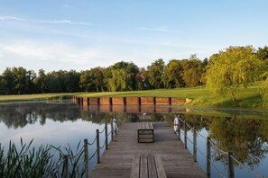 Eine hölzerne Mole, die sich über einen stillen See erstreckt, mit einer grasbewachsenen Fläche und Bäumen im Hintergrund.