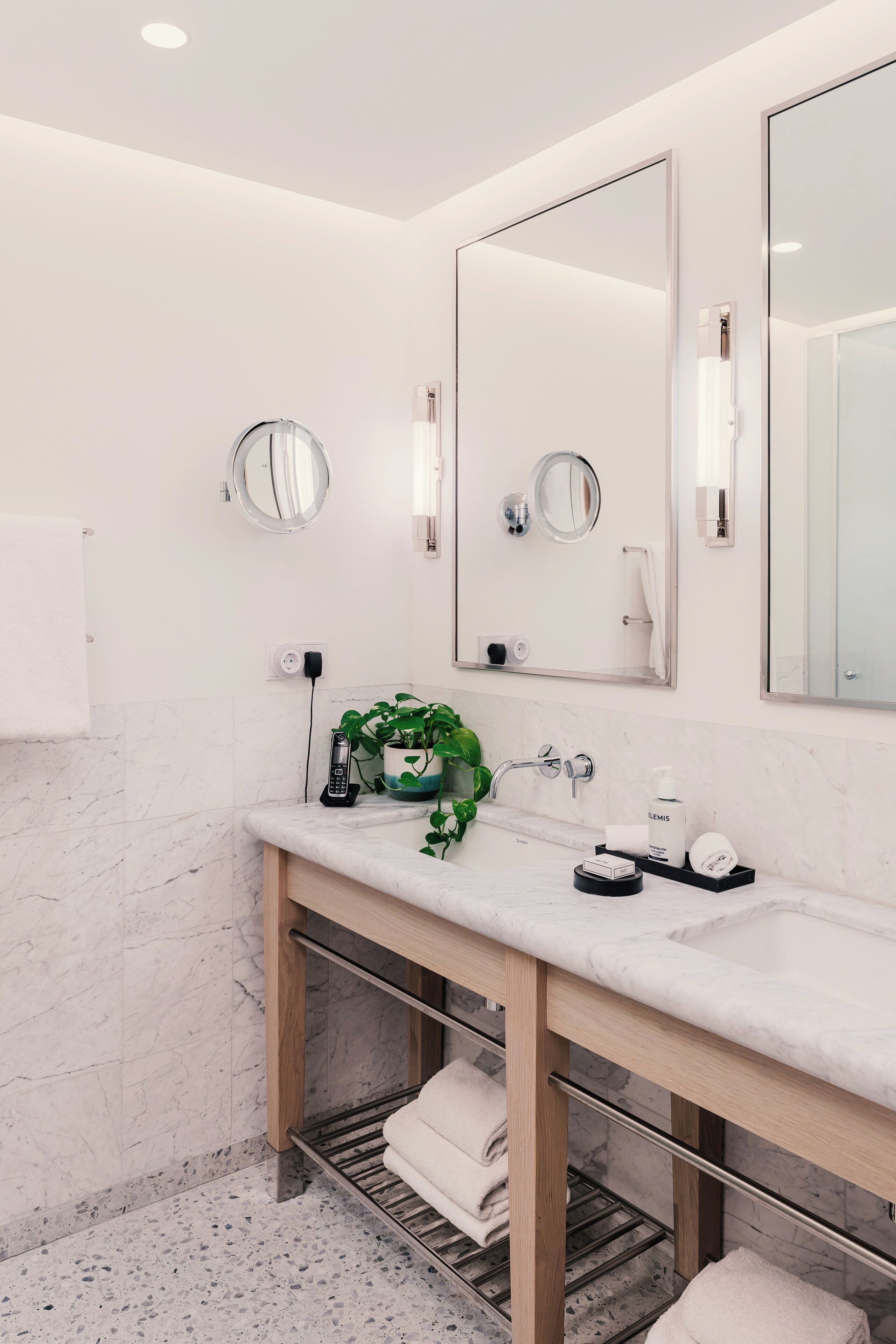A modern bathroom with a marble sink, two mirrors, a potted plant, and a towel rack.