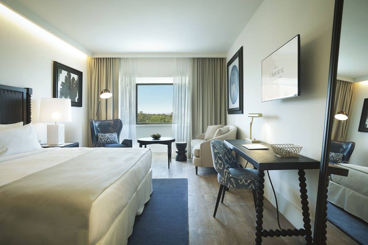 Une chambre d’hôtel avec un grand lit, deux fauteuils, un bureau avec une lampe, une petite table et une vue sur l’extérieur.