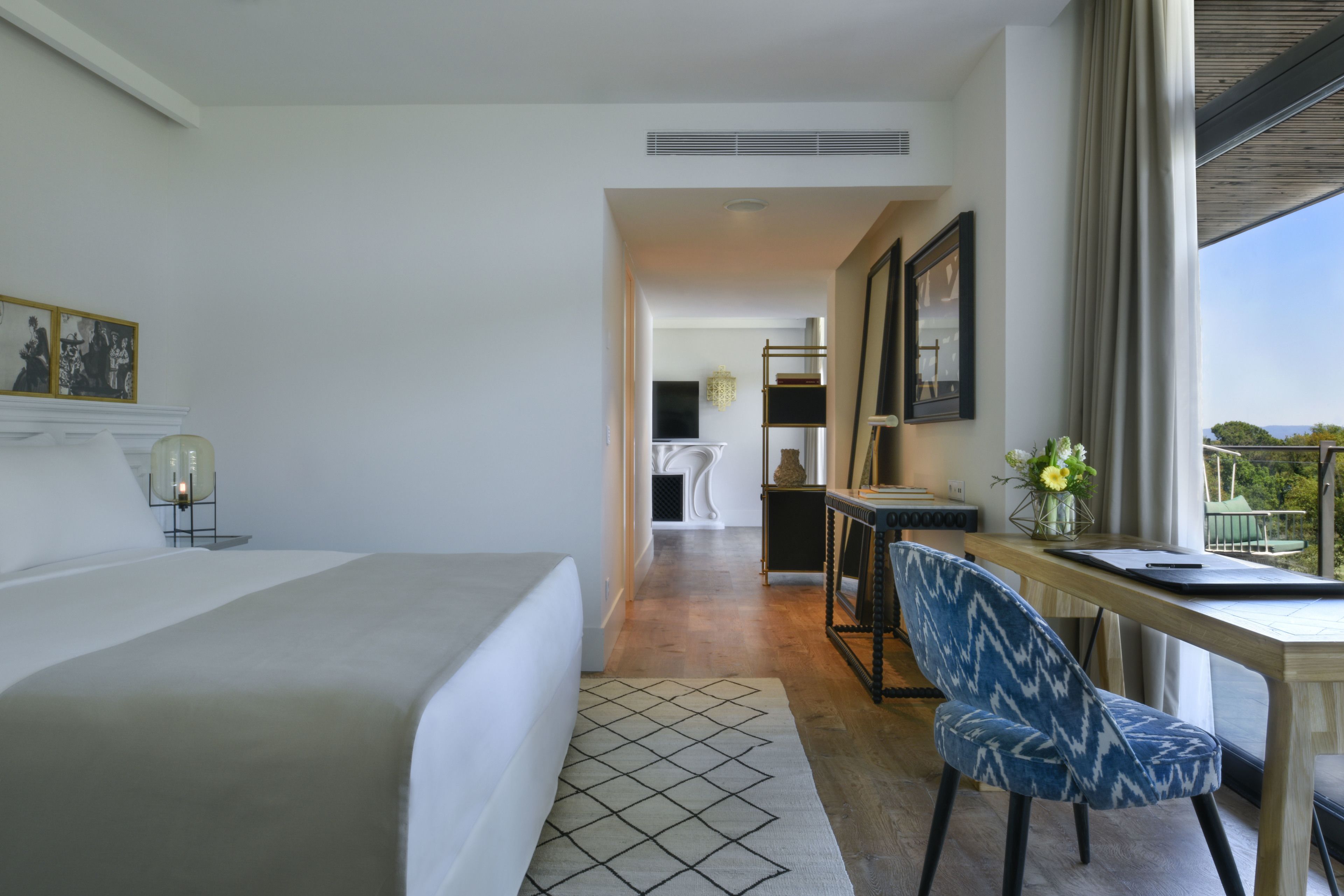 A hotel room with a large bed, a desk with a chair, a view of the outdoors, and a small table with a vase of flowers.