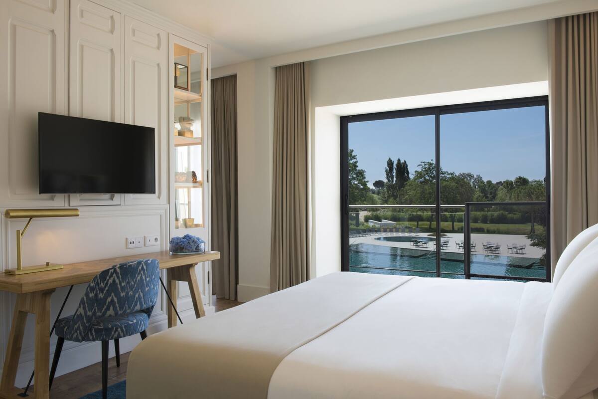 Une chambre d’hôtel avec un lit, un bureau avec une télévision, une chaise, une fenêtre donnant sur une piscine et un espace verdoyant.