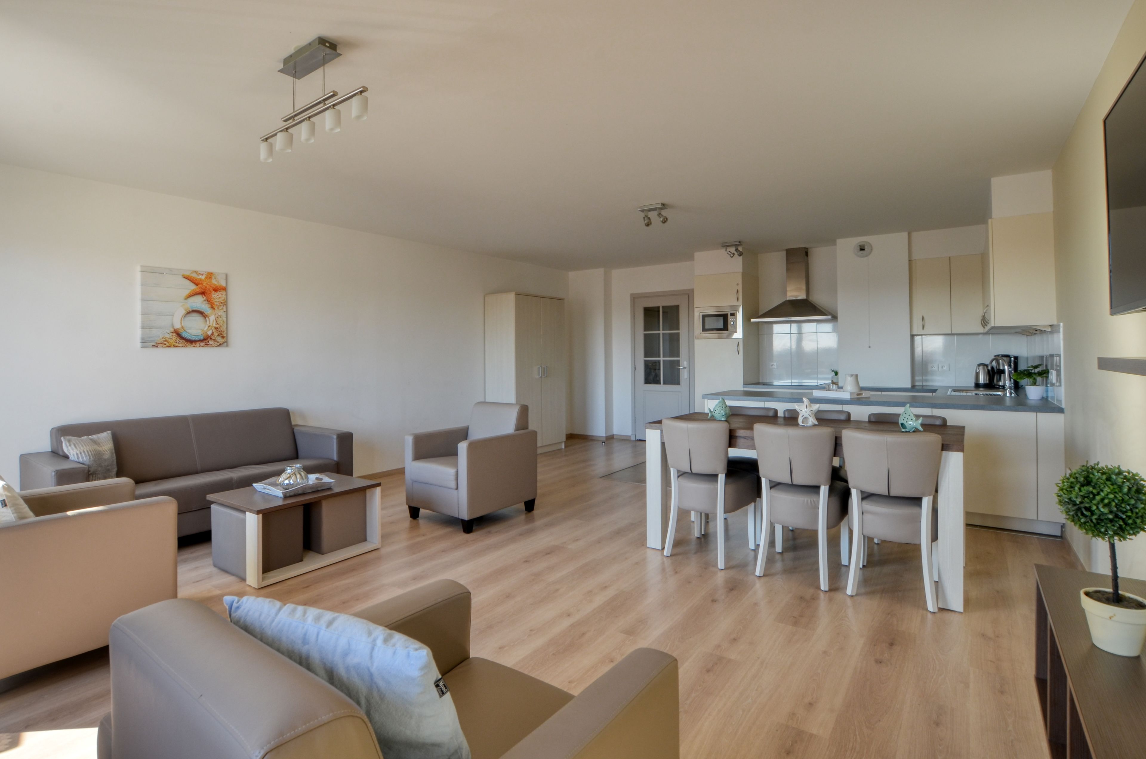 A modern living room with a sofa, armchairs, and a dining area with a table and chairs. A kitchen is visible in the background with cabinets and a microwave.