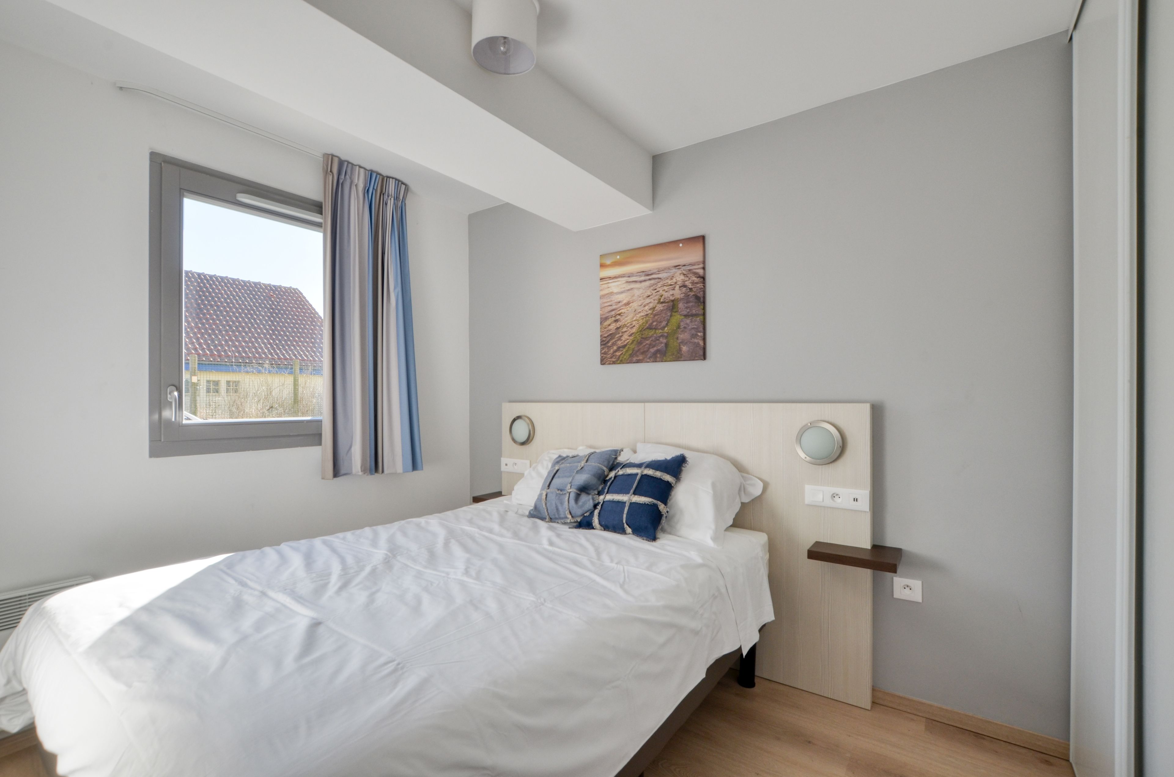 Un dormitori modern amb un llit gran, una finestra amb cortines, una làmpada de paret i un quadre.