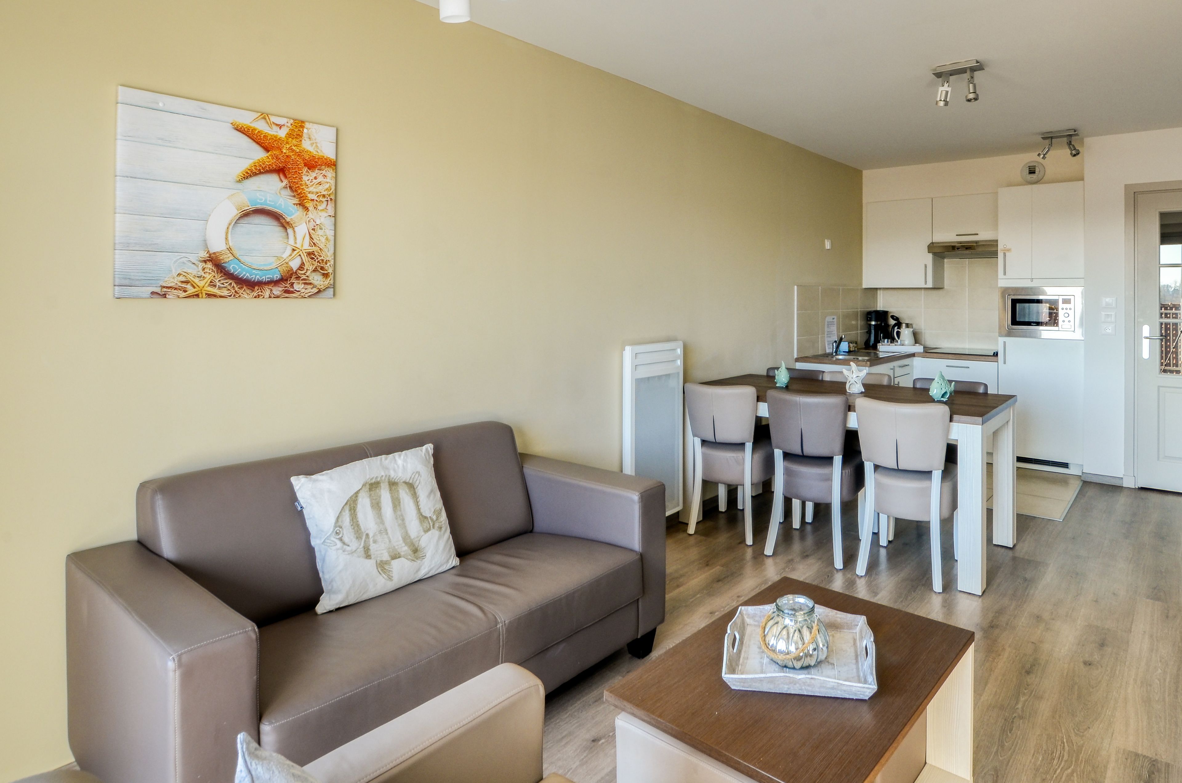 Uma sala de estar moderna com um sofá marrom, uma mesa de centro e uma área de jantar com cadeiras, além de uma pequena cozinha ao fundo.