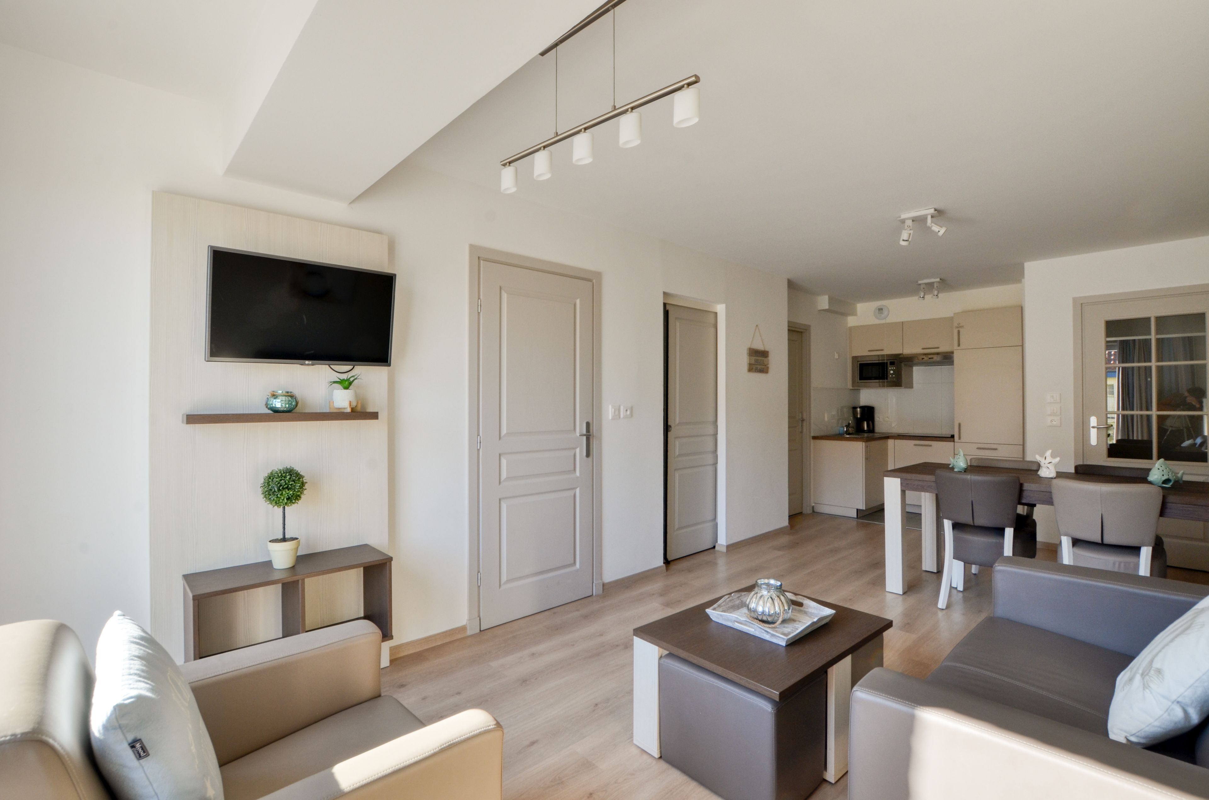 A modern living room with a flat-screen TV, a coffee table, and a sofa. There is a kitchen area with a dining table and chairs.