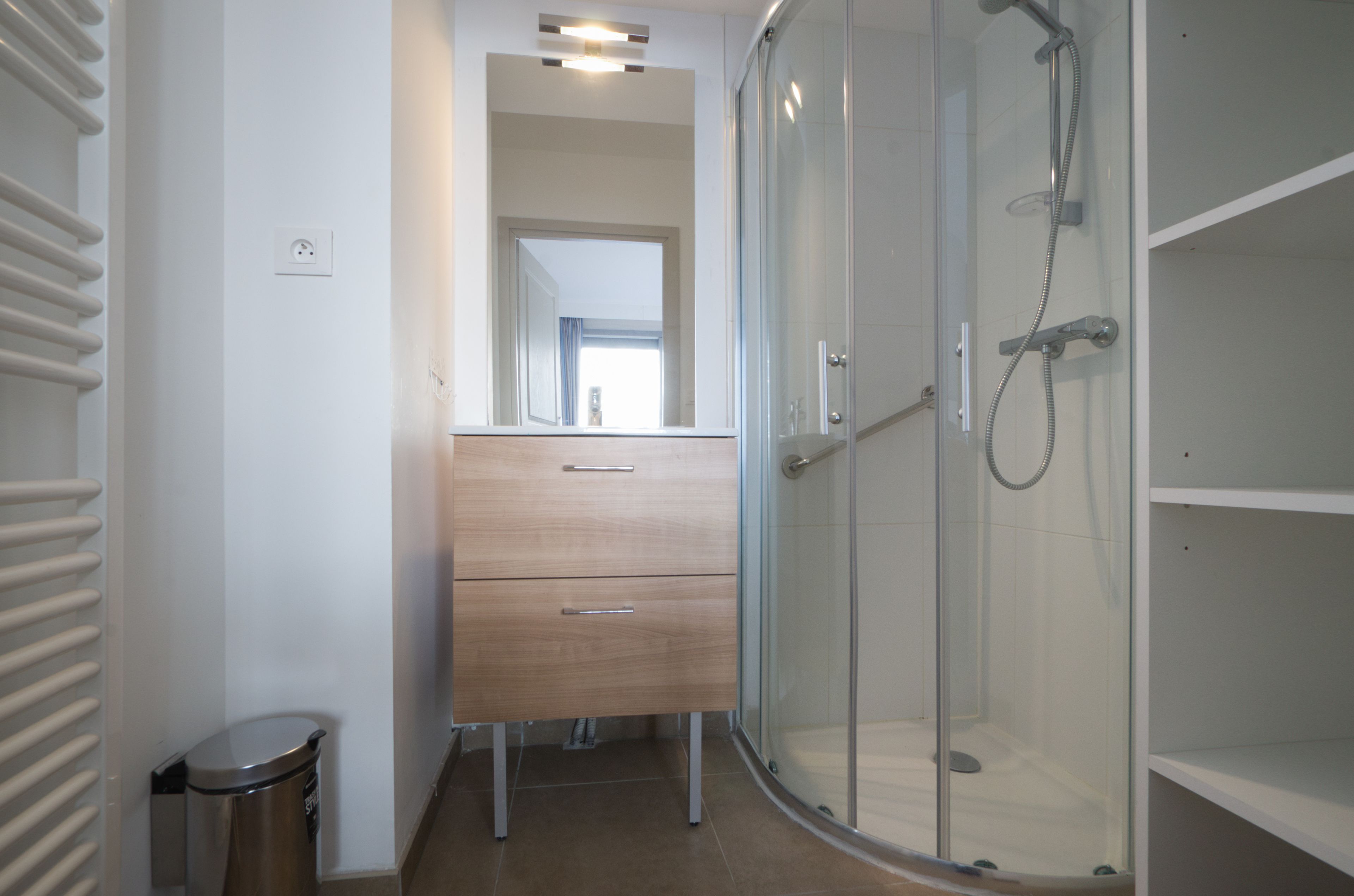 A modern bathroom with a glass shower enclosure, a wooden vanity with drawers, and a trash bin.