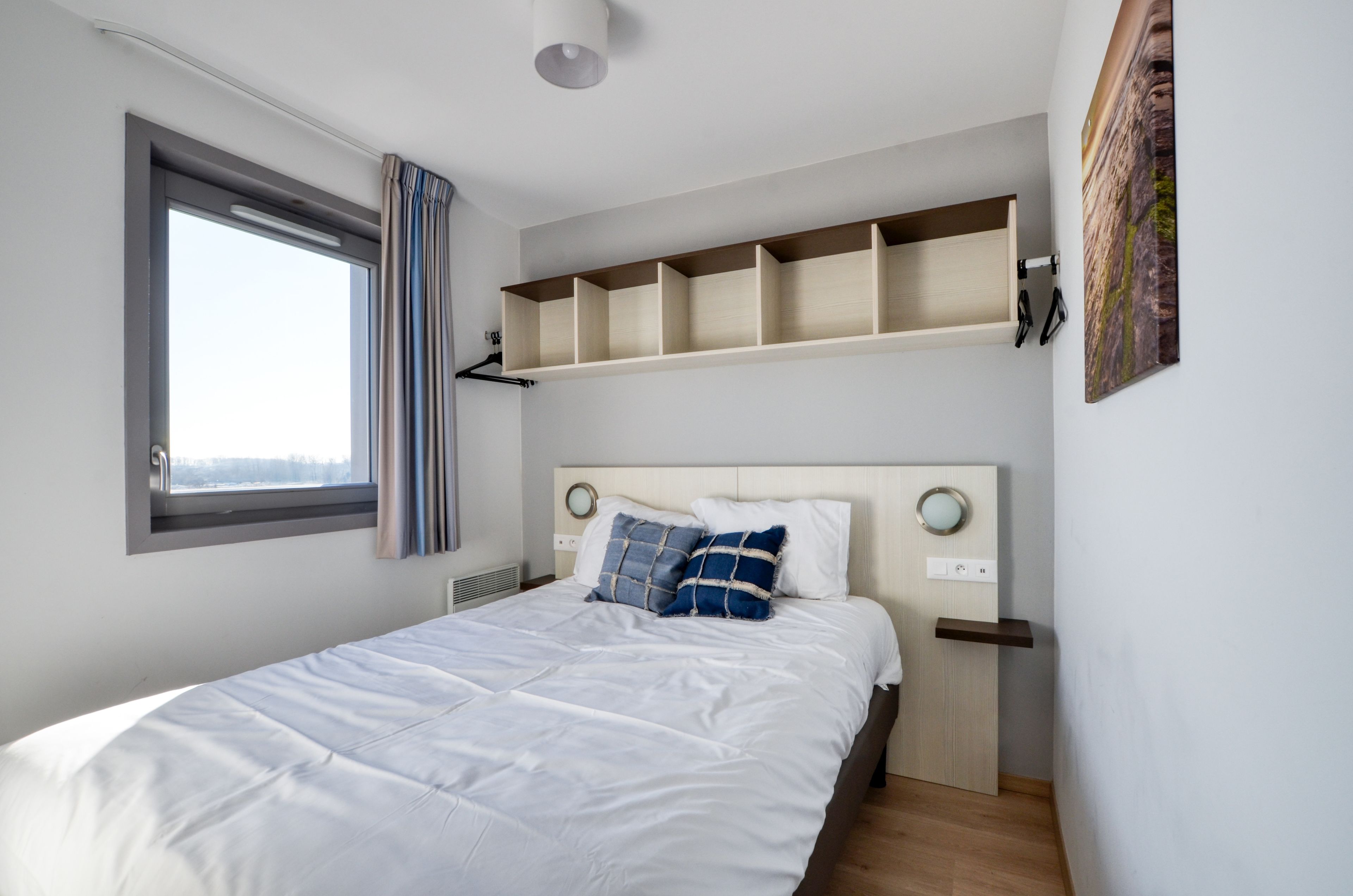 Ein Schlafzimmer mit einem Bett, einem Fenster mit Vorhängen, einer eingebauten Ablage über dem Bett und einer Wandleuchte.