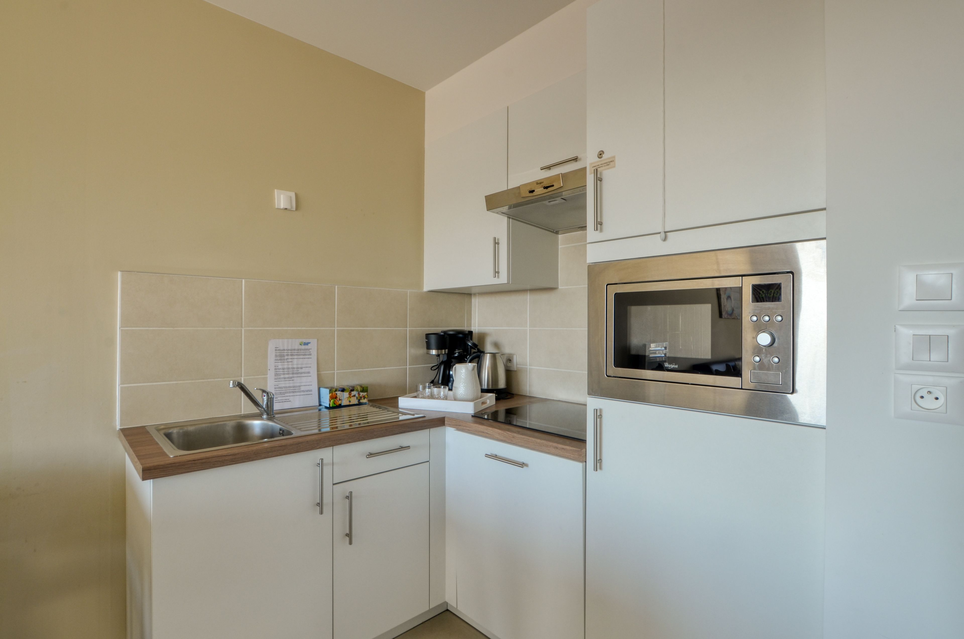 Una cocina compacta con armarios blancos, un microondas de acero inoxidable, una cafetera y un lavamanos.