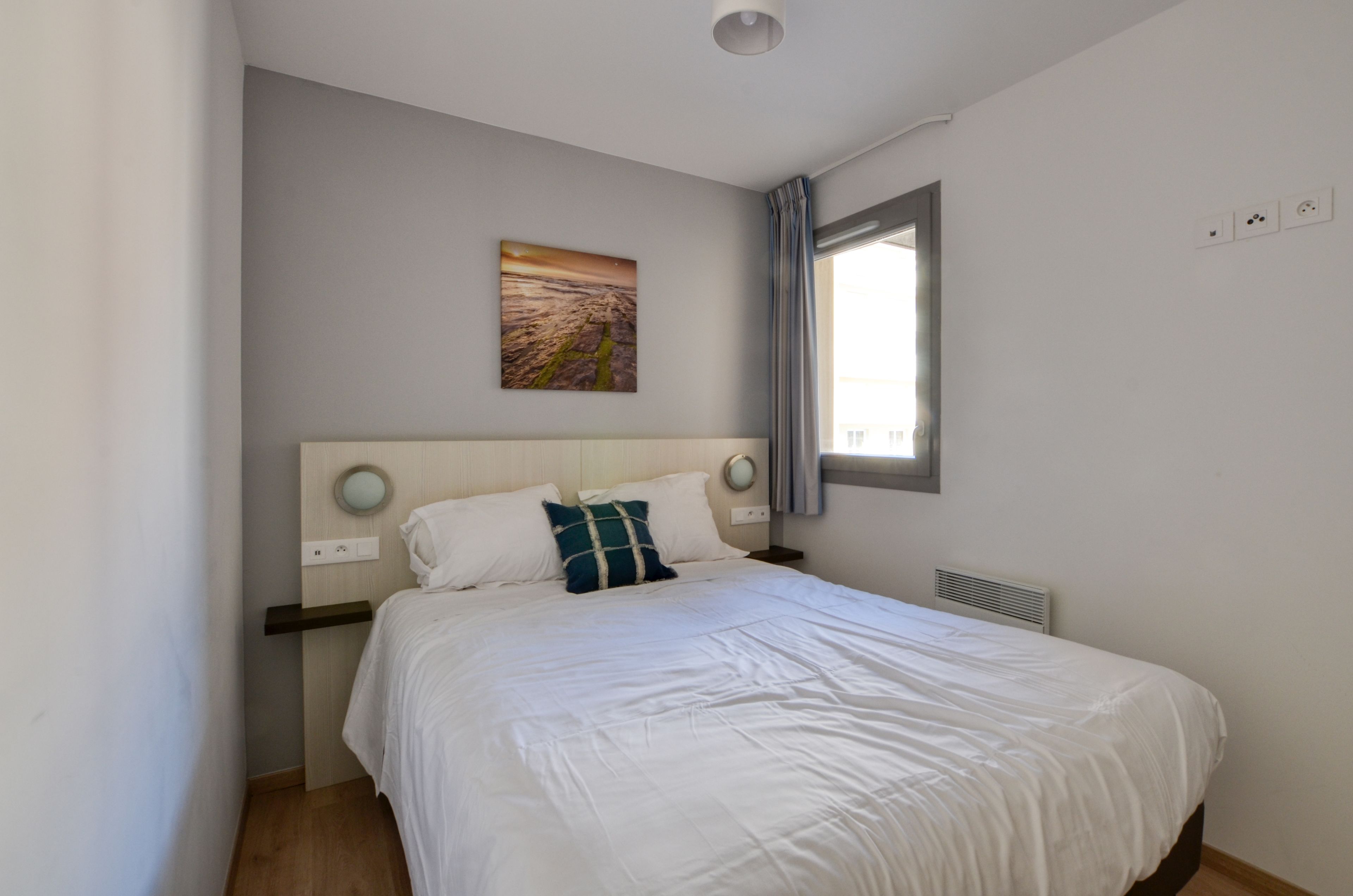 A neatly made bed with white bedding and a single teal pillow, a wall-mounted picture, a window with curtains, and a bedside table with a lamp.
