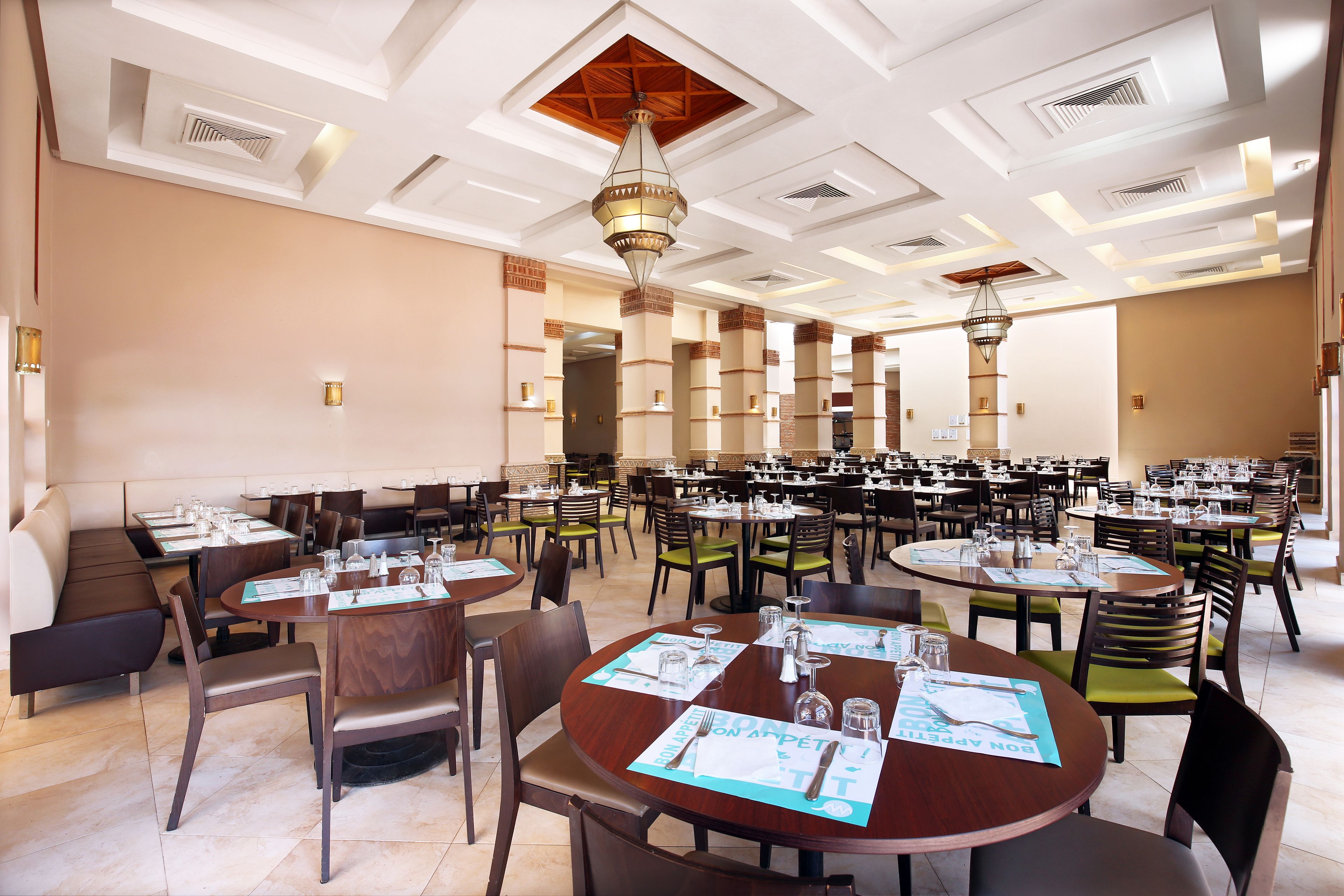 A spacious dining area with multiple tables and booths, featuring a high ceiling with recessed lighting and a central chandelier.