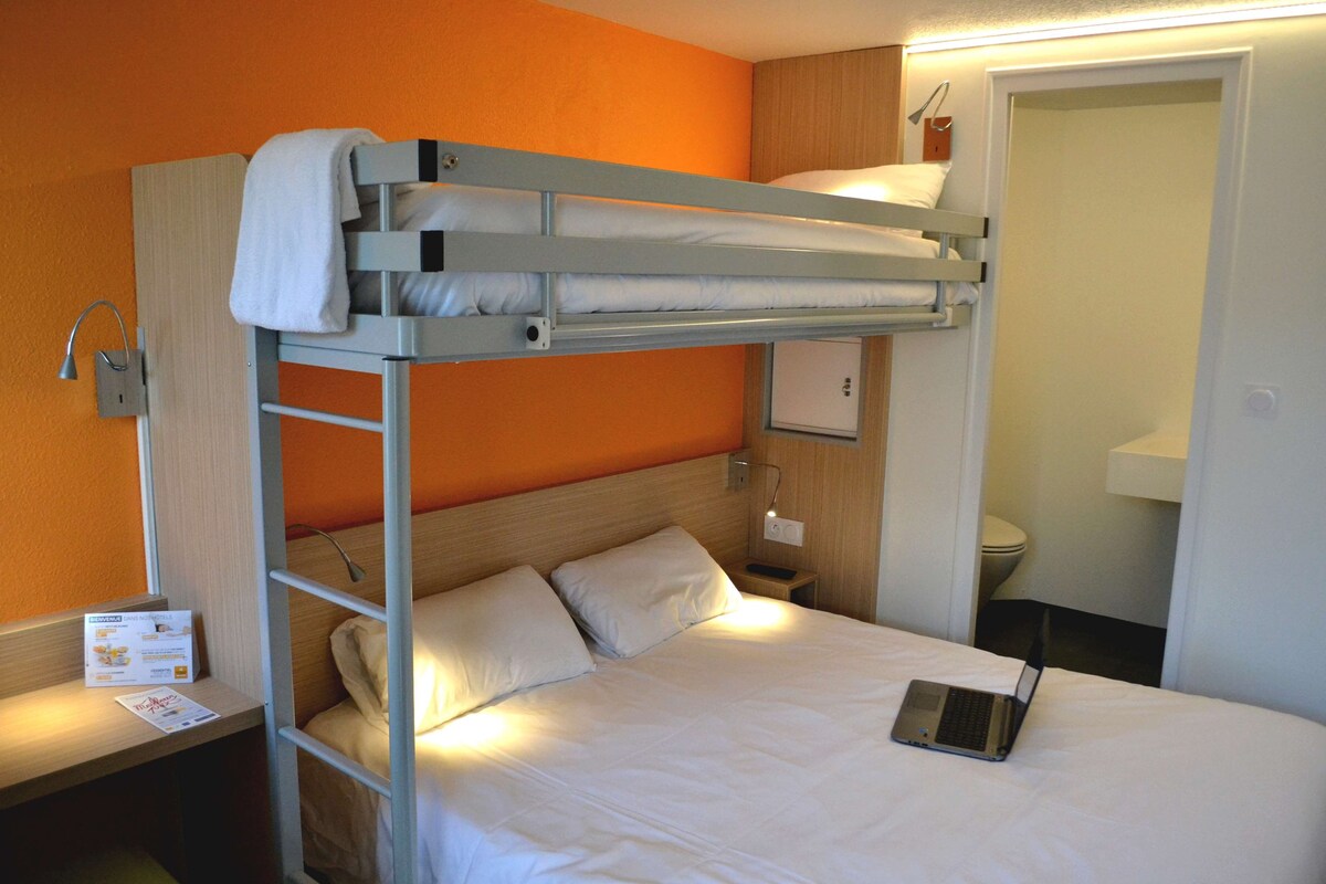 A bunk bed with a laptop on the lower bunk, a bedside table with a lamp, and a small nightstand with a sign.