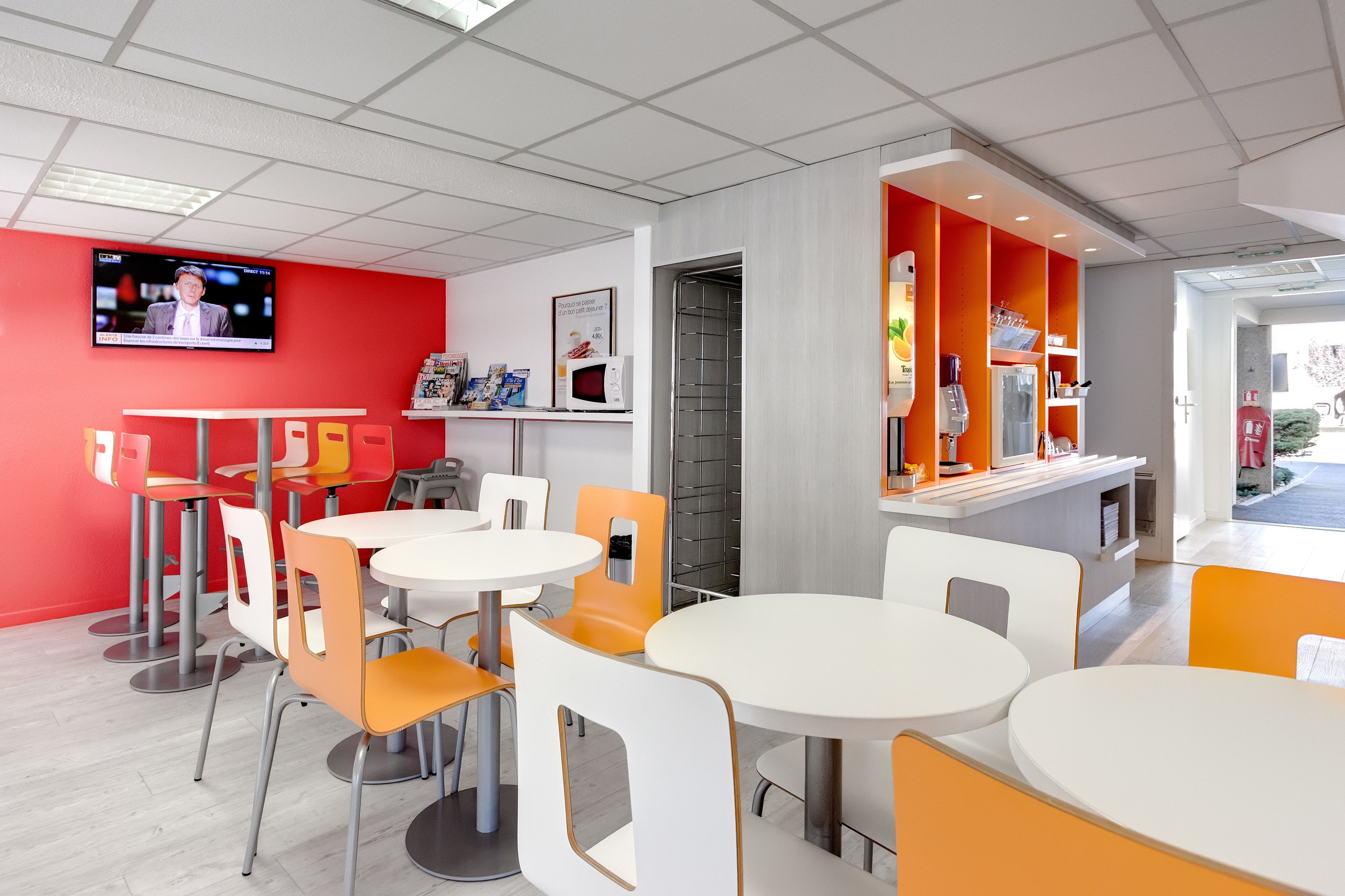 Un restaurante moderno con mesas y sillas blancas y anaranjadas, una pared de acento roja y una pantalla de televisión que muestra un noticiero.