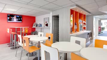 Un restaurante moderno con mesas y sillas blancas y anaranjadas, una pared de acento roja y una pantalla de televisión que muestra un noticiero.