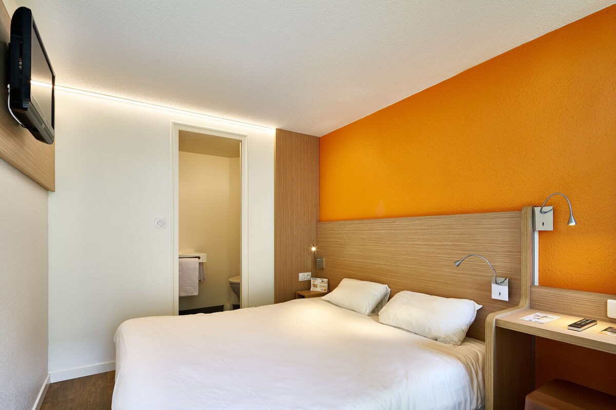 A hotel room with a bed, bedside table, lamp, and a TV mounted on the wall.