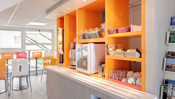 A modern cafe with a bright orange color scheme, featuring a coffee machine, seating area, and various storage containers. - Première Classe La Roche Sur Yon - Vendéspace (Mouilleron-le-Captif)