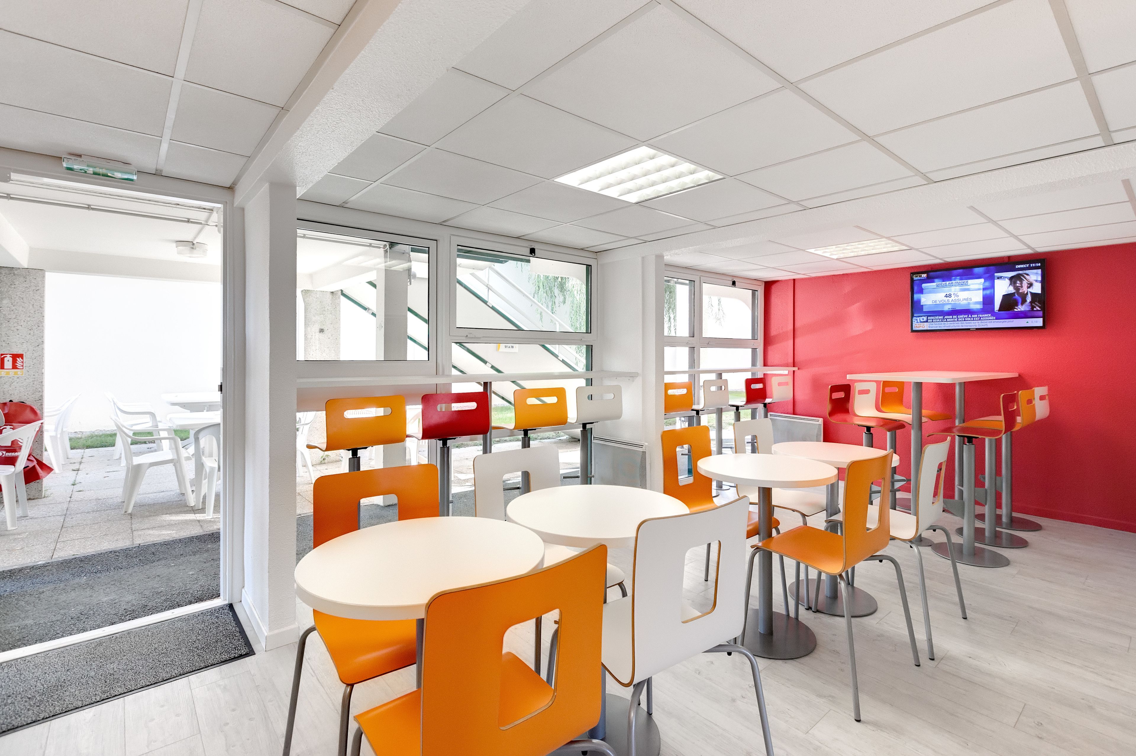Un comedor moderno con sillas blancas y anaranjadas, mesas redondas y una pared roja con una pantalla de televisión.