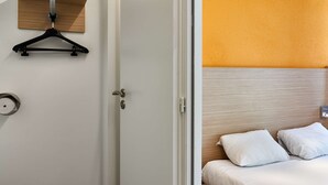 A hotel room with a bed, a wooden headboard, a white wall, a wooden shelf with a black hanger, and a door leading to another room. - Première Classe La Roche Sur Yon - Vendéspace (Mouilleron-le-Captif)