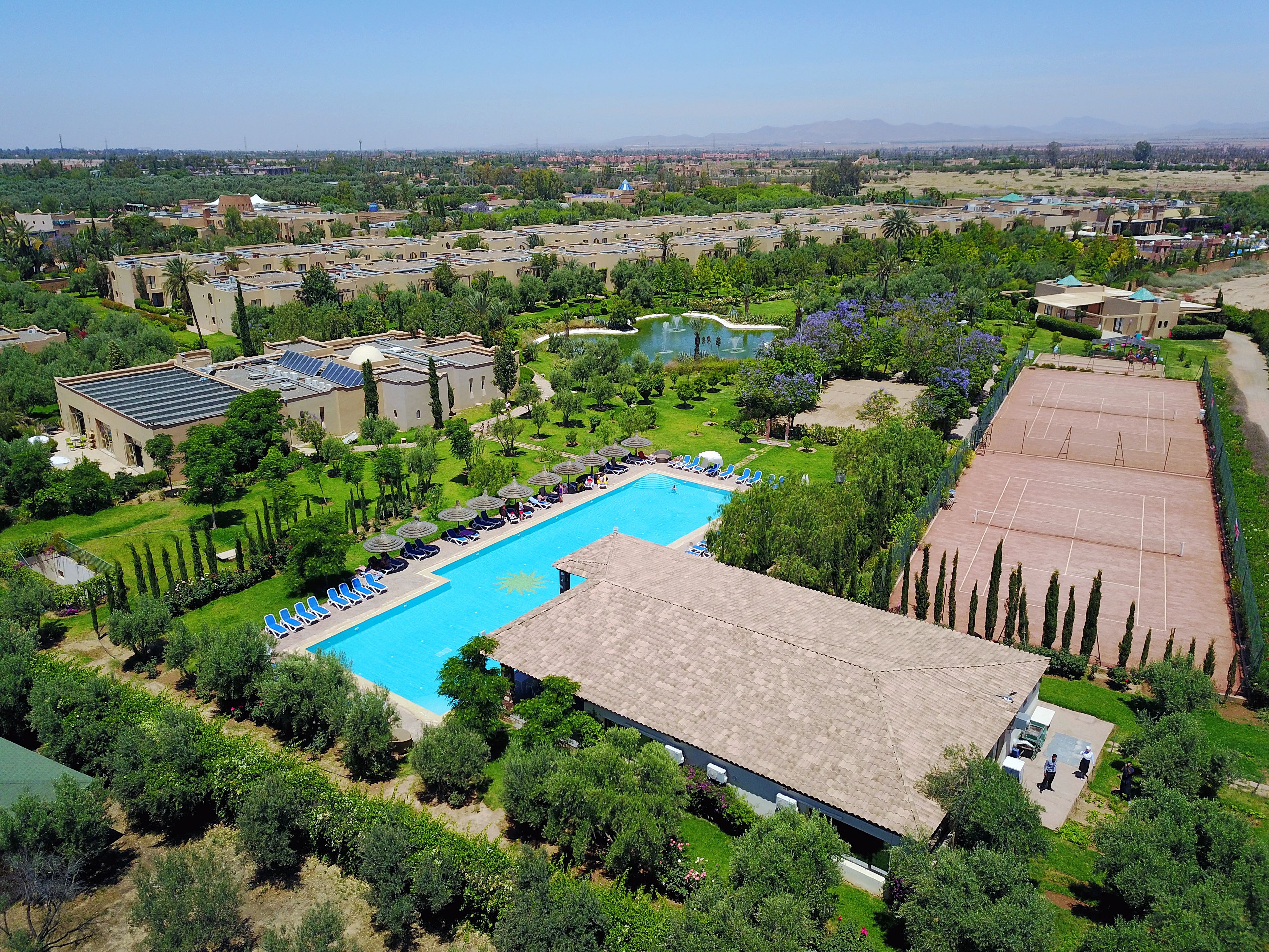 An aerial view of a resort with a swimming pool, sun loungers, and a tennis court.
