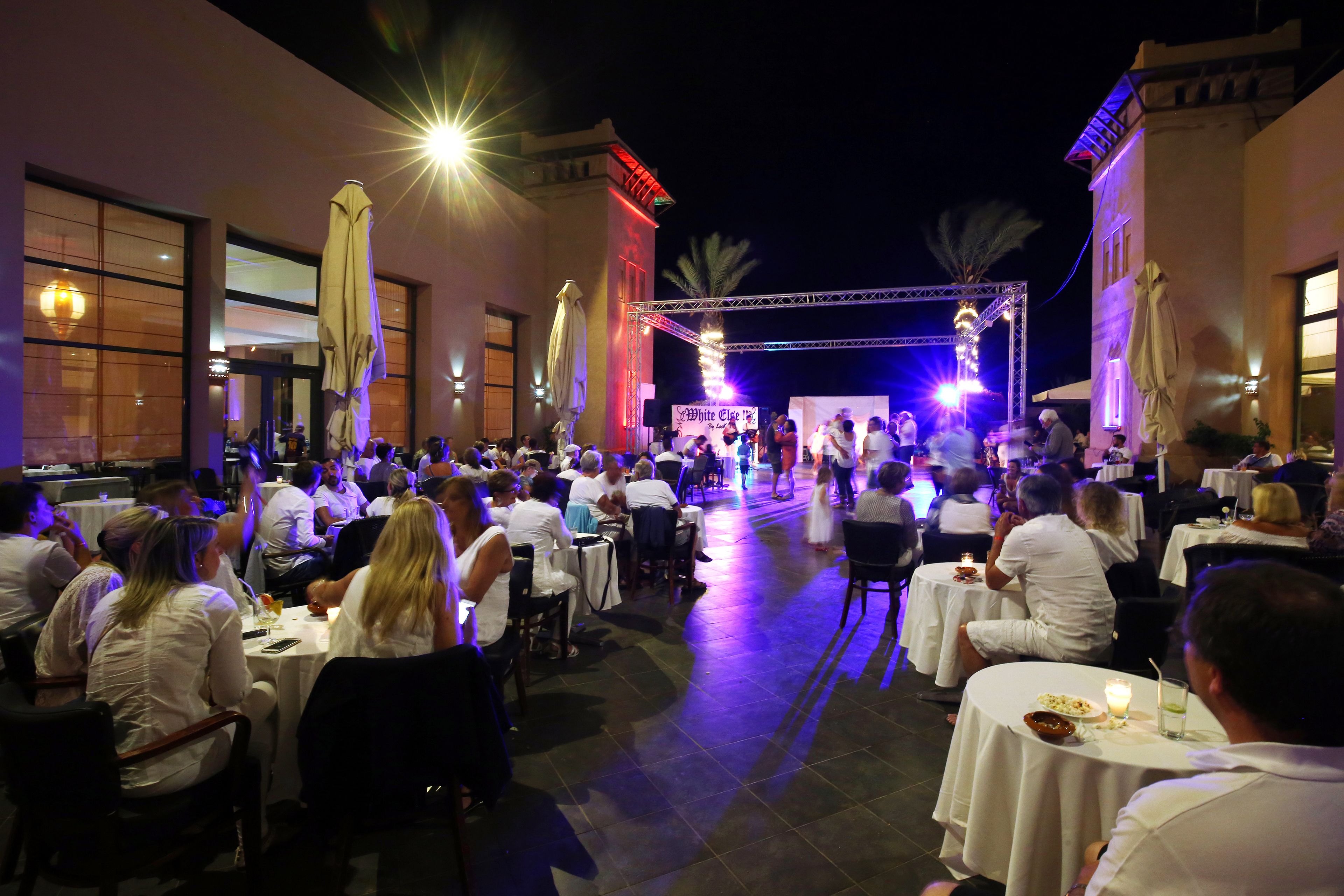An outdoor dining area with round tables and umbrellas, surrounded by buildings with large windows and a stage in the background.