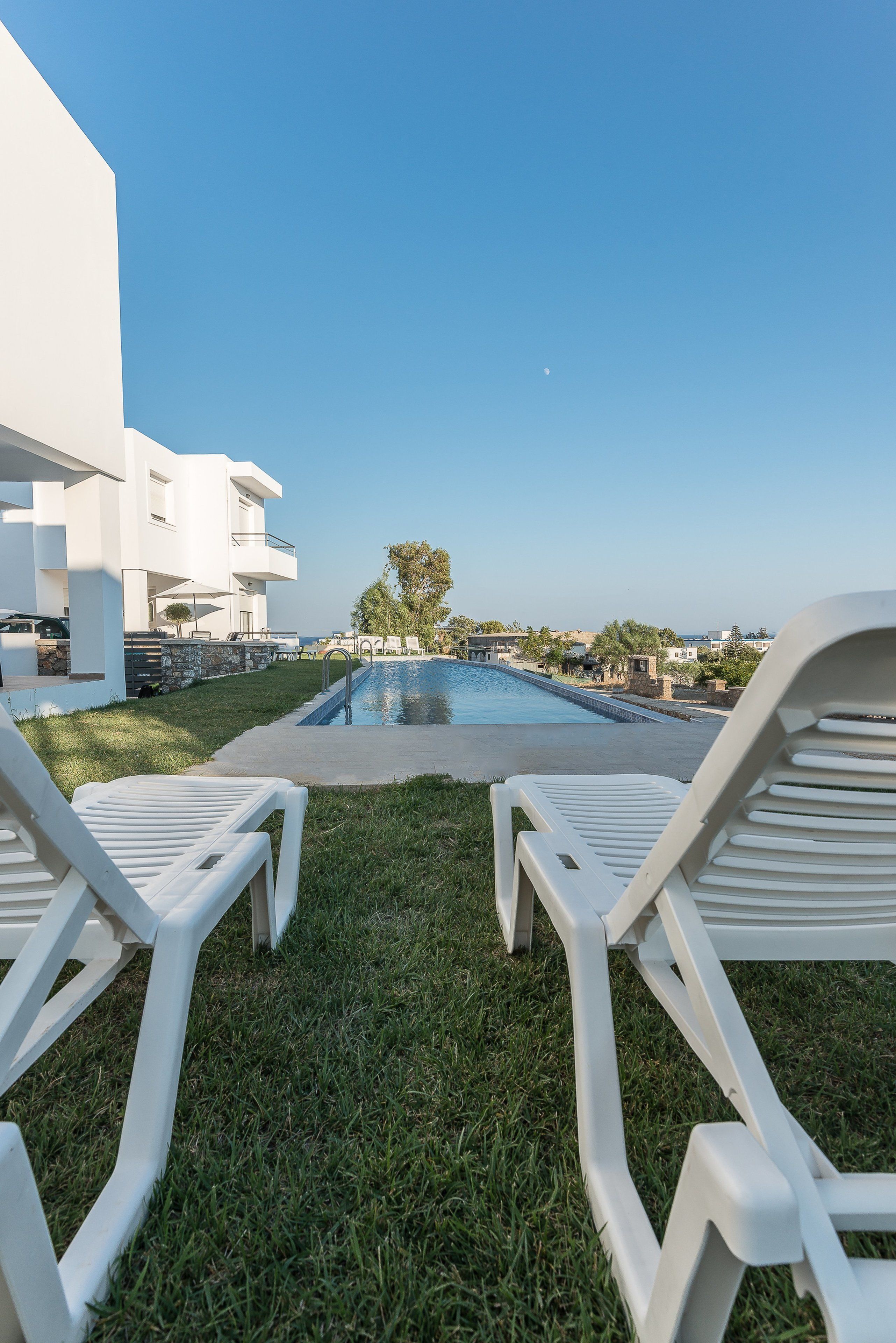 A pool area with two white lounge chairs facing the pool, surrounded by a grassy lawn and a white building.