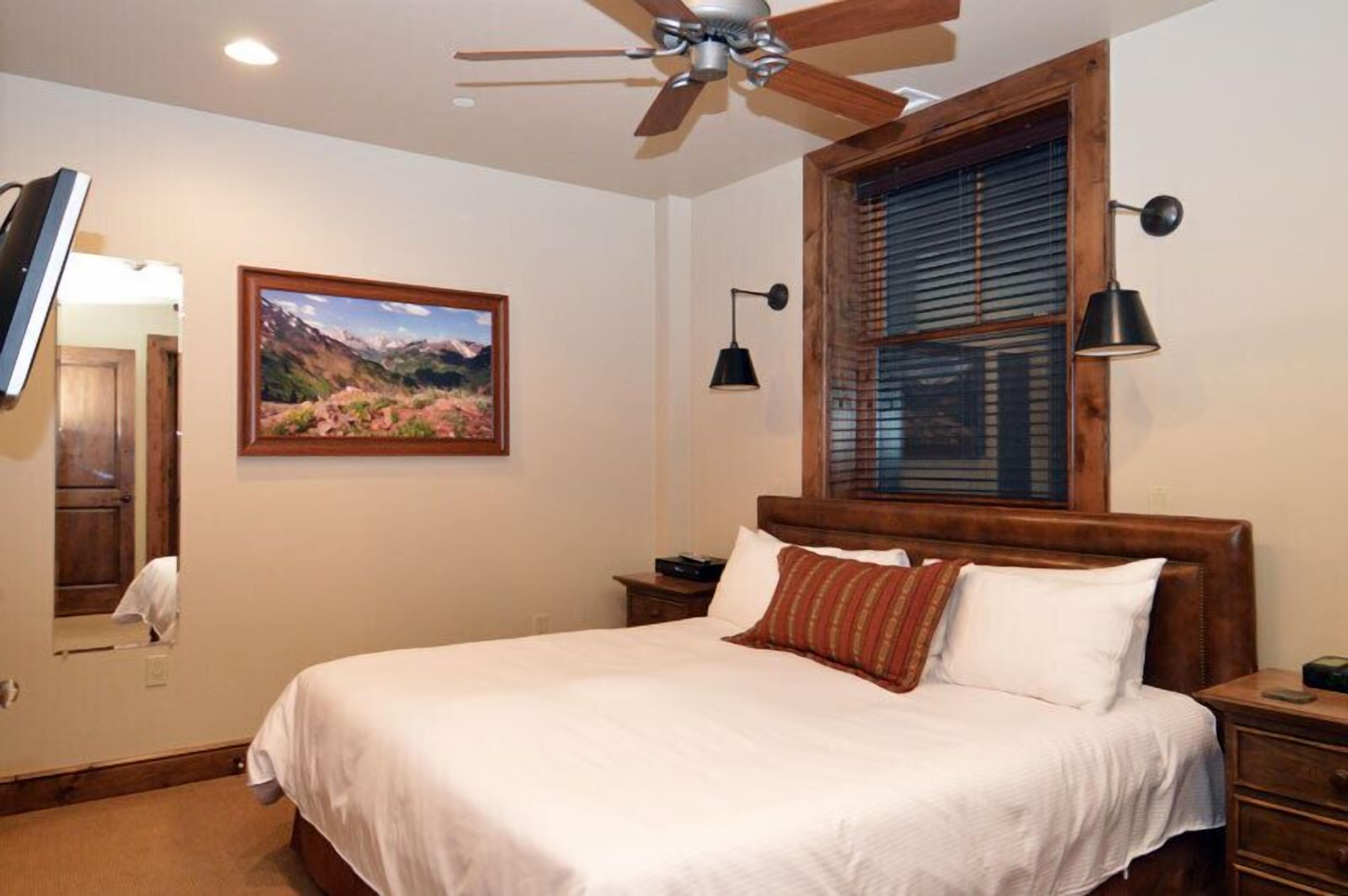 A bedroom with a large bed, a ceiling fan, a window with blinds, a mounted TV, and a framed picture on the wall.
