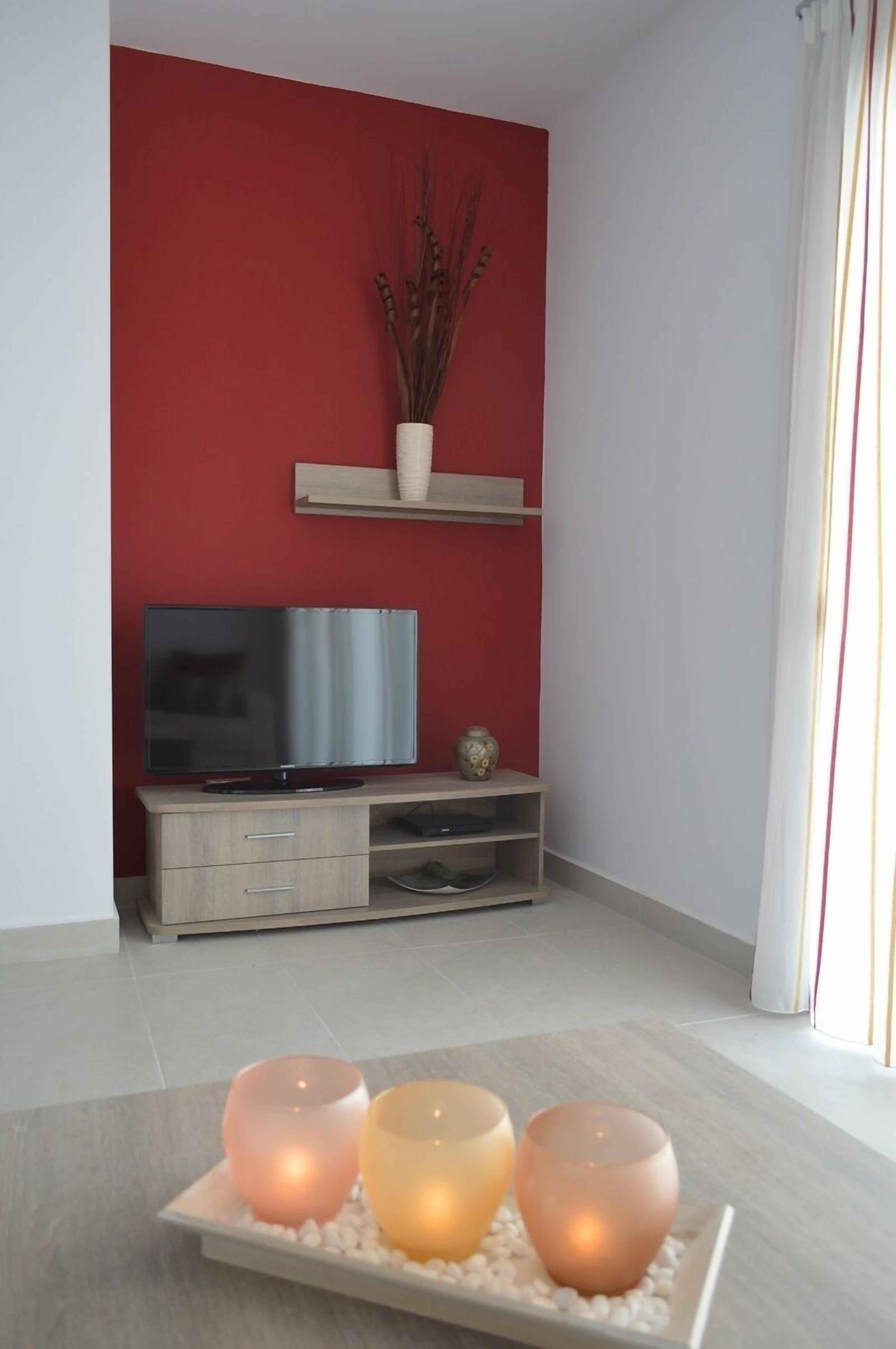 Un salon moderne avec un mur d’accent rouge, un téléviseur à écran plat posé sur un meuble en bois et un vase décoratif sur une étagère.