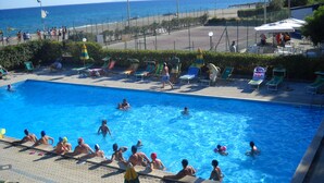 A swimming pool with people in and around it, a beach in the background, and lounge chairs and umbrellas on the poolside.
