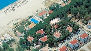 An aerial view of a coastal area with a beach, a tennis court, and a cluster of buildings with red roofs.