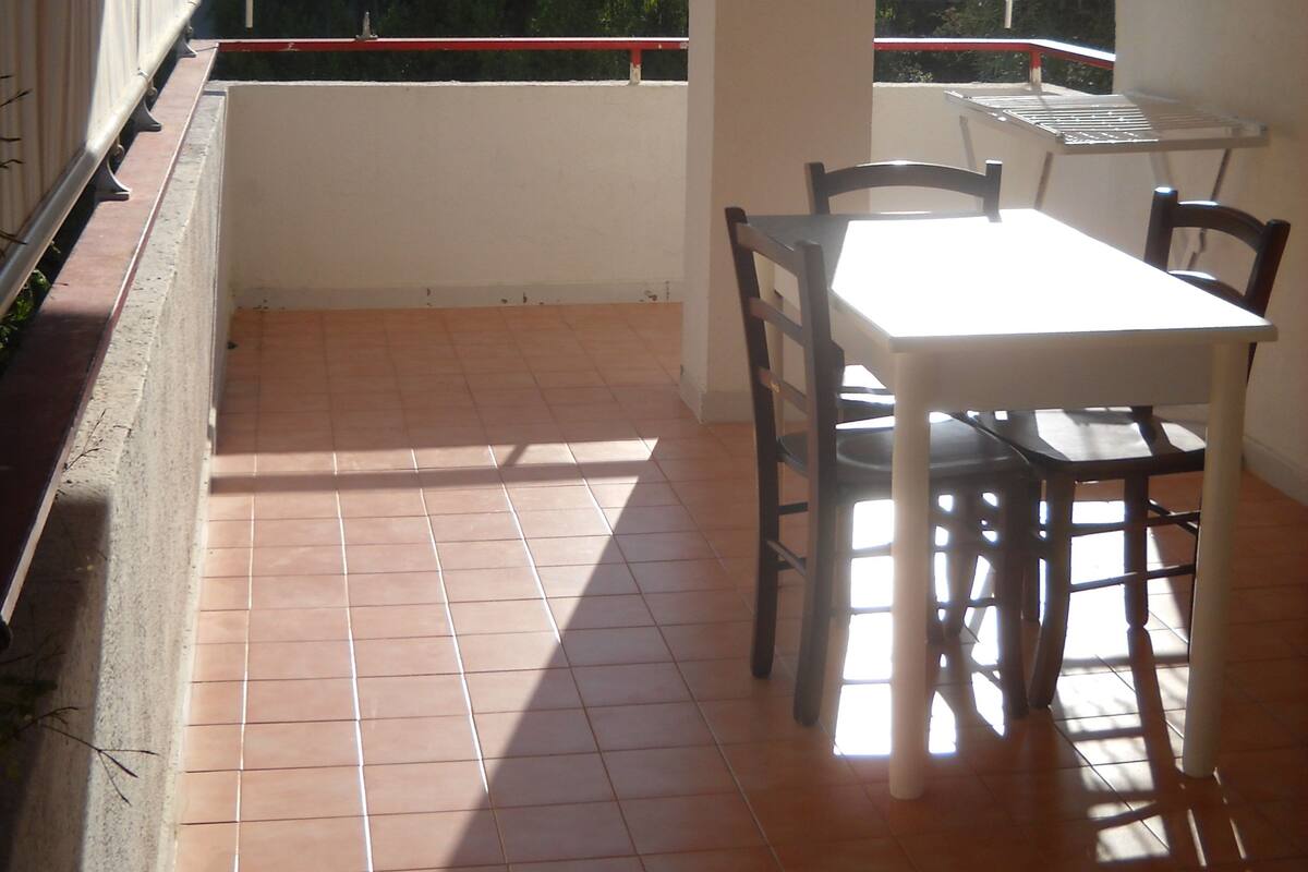 Een balkon met betegelde vloer, een witte tafel met stoelen en uitzicht op bomen en gebouwen.