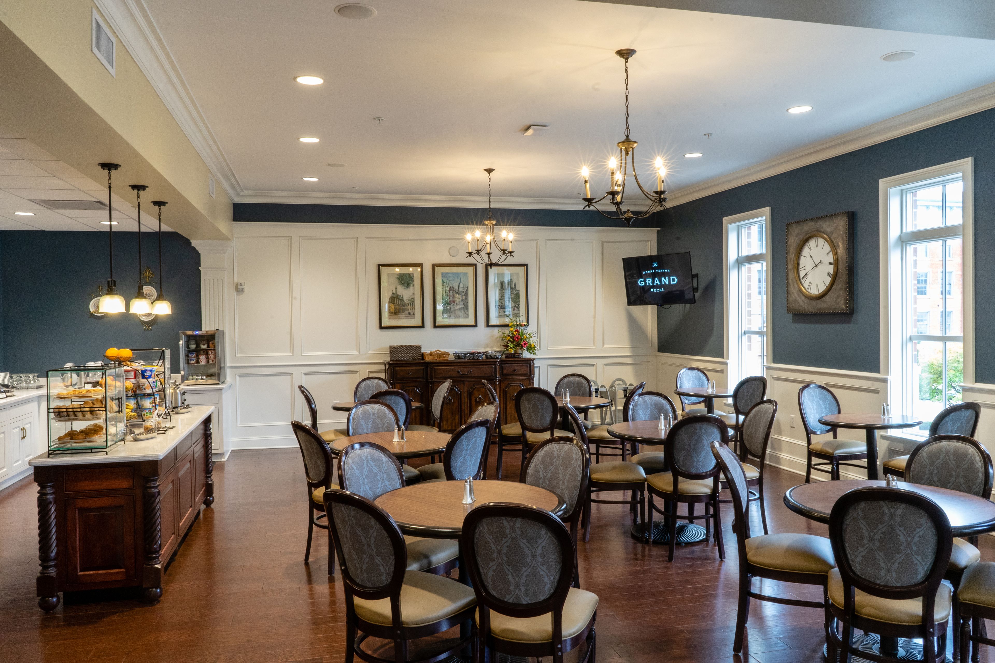 A spacious dining area with multiple round tables and chairs, a buffet counter with food displays, and a television mounted on the wall.