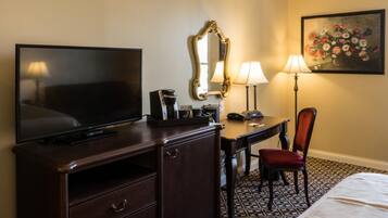 A hotel room with a TV, a desk with a lamp, a chair, a dresser, and a painting on the wall.