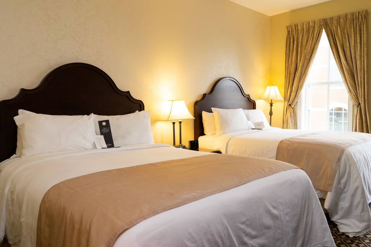 Une chambre d’hôtel avec deux lits, chacun doté d’une tête de lit en bois, de draps blancs et d’un plaid beige. On y trouve des lampes de chevet et une fenêtre avec des rideaux.