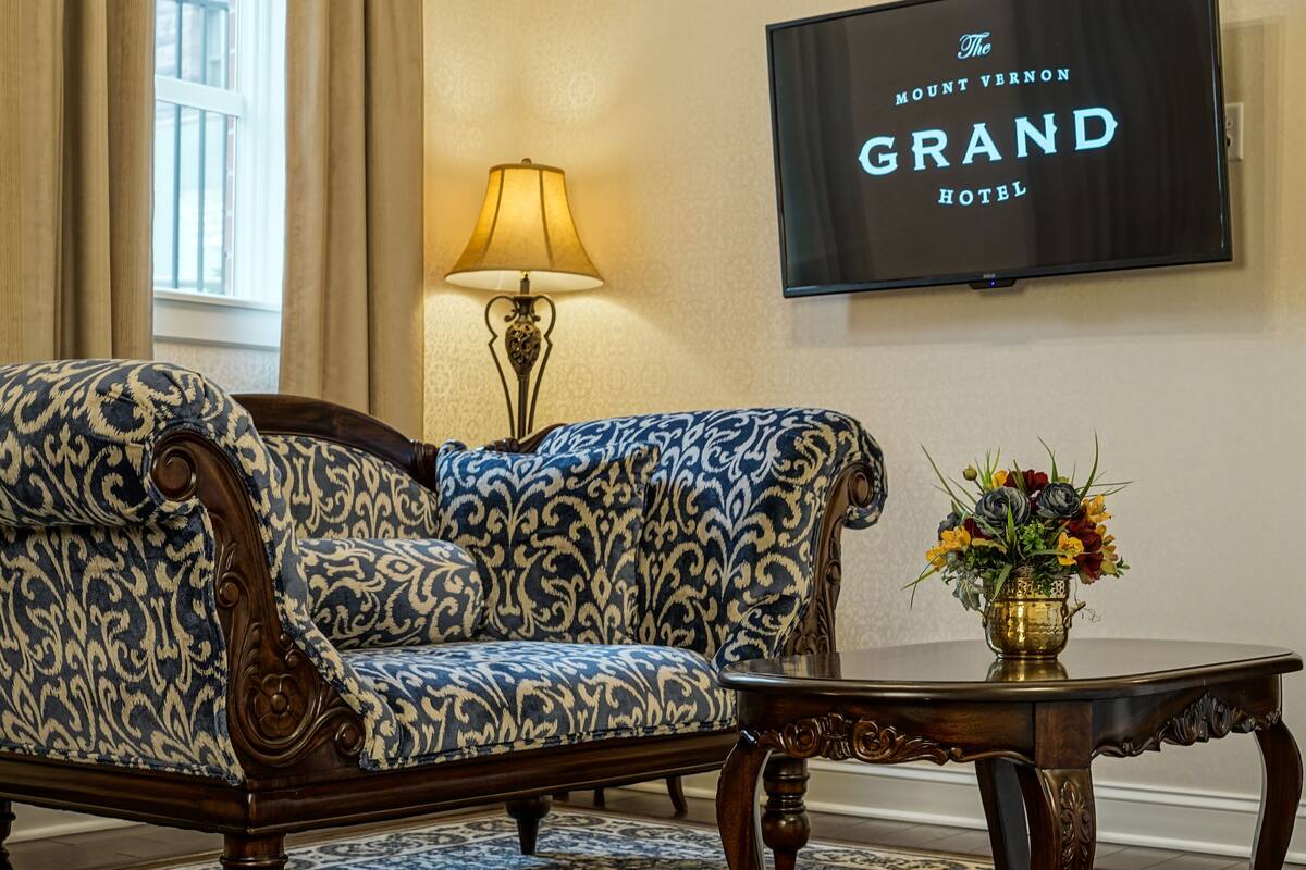 Une pièce avec un canapé à motifs, une table ornée d’un vase de fleurs et un téléviseur fixé au mur qui affiche l’enseigne « The Mount Vernon Grand Hotel ».