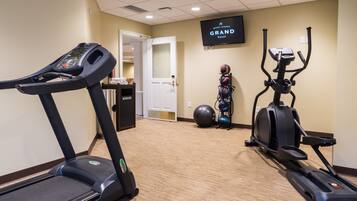 A hotel fitness room with a treadmill, elliptical, stationary bike, and a large exercise ball. A television screen displays the hotel name.