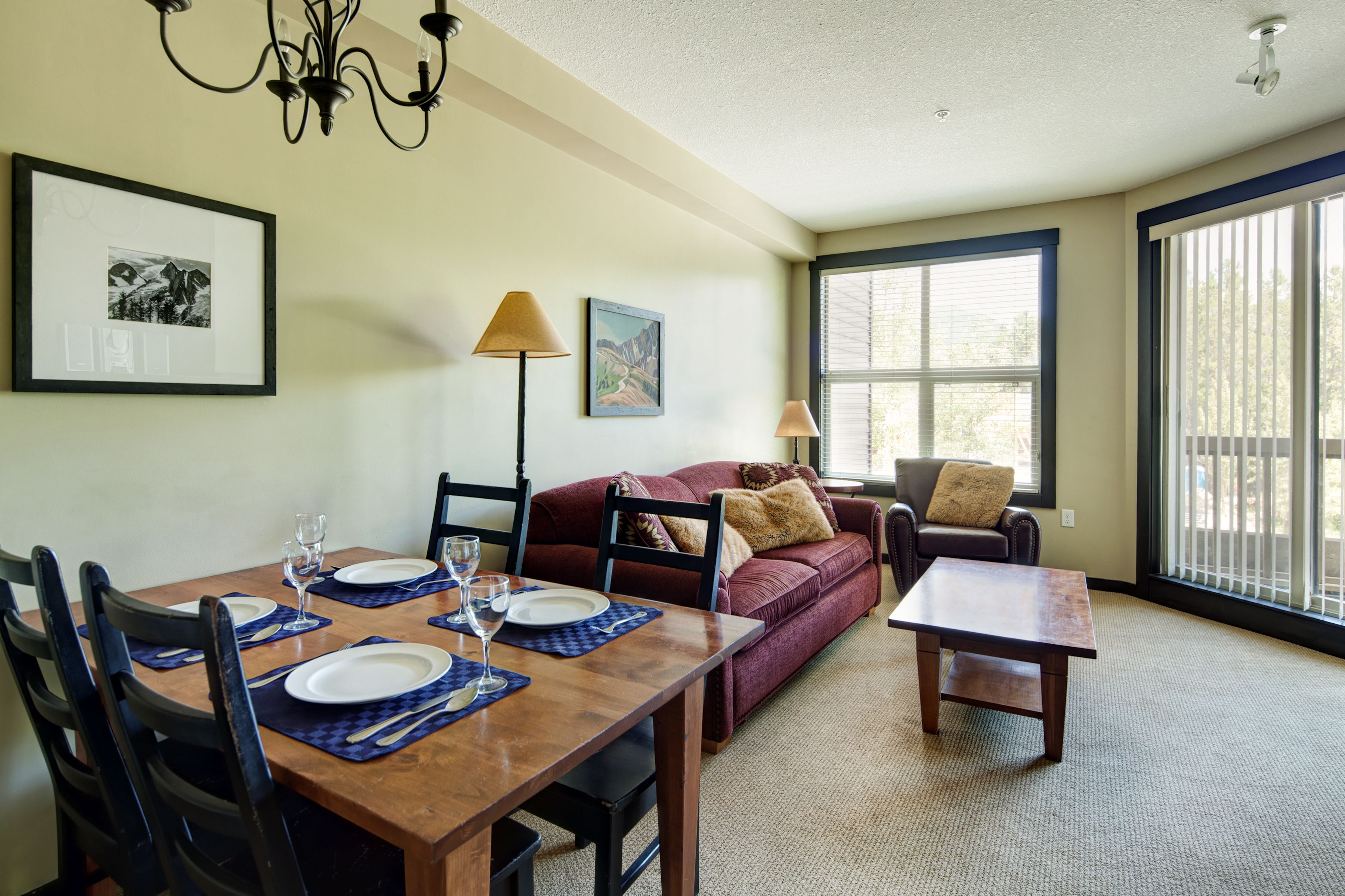 A dining area with a wooden table set for four, a red sofa, and a window with blinds.