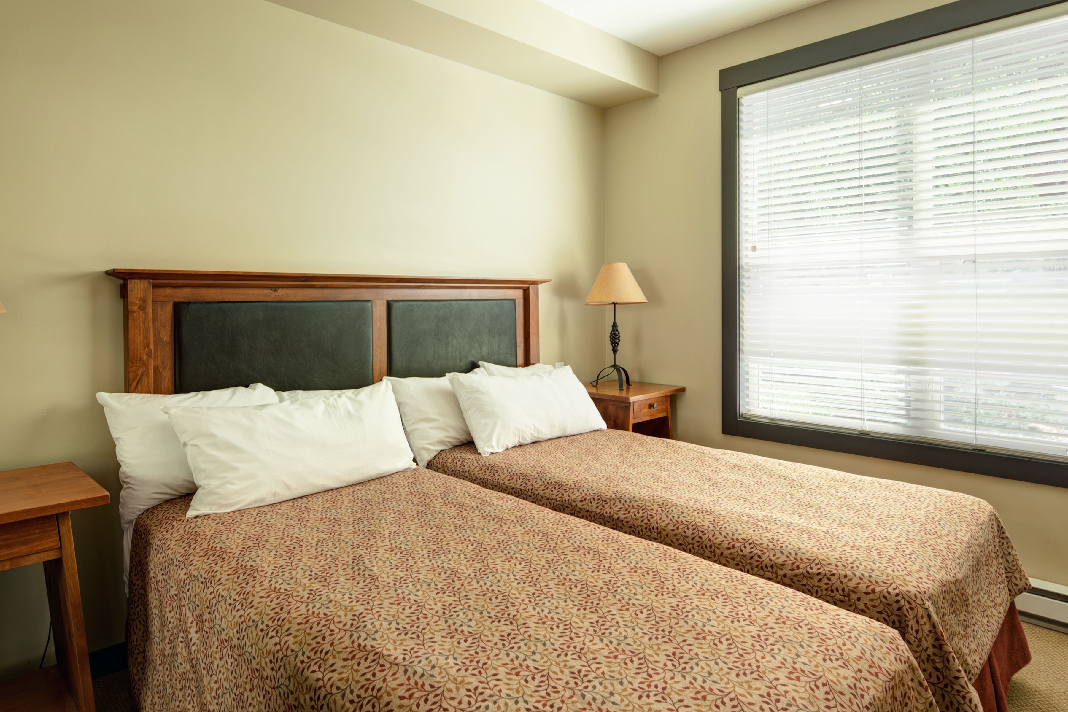 A double bed with a wooden headboard, two bedside tables, a lamp, and a large window with blinds.