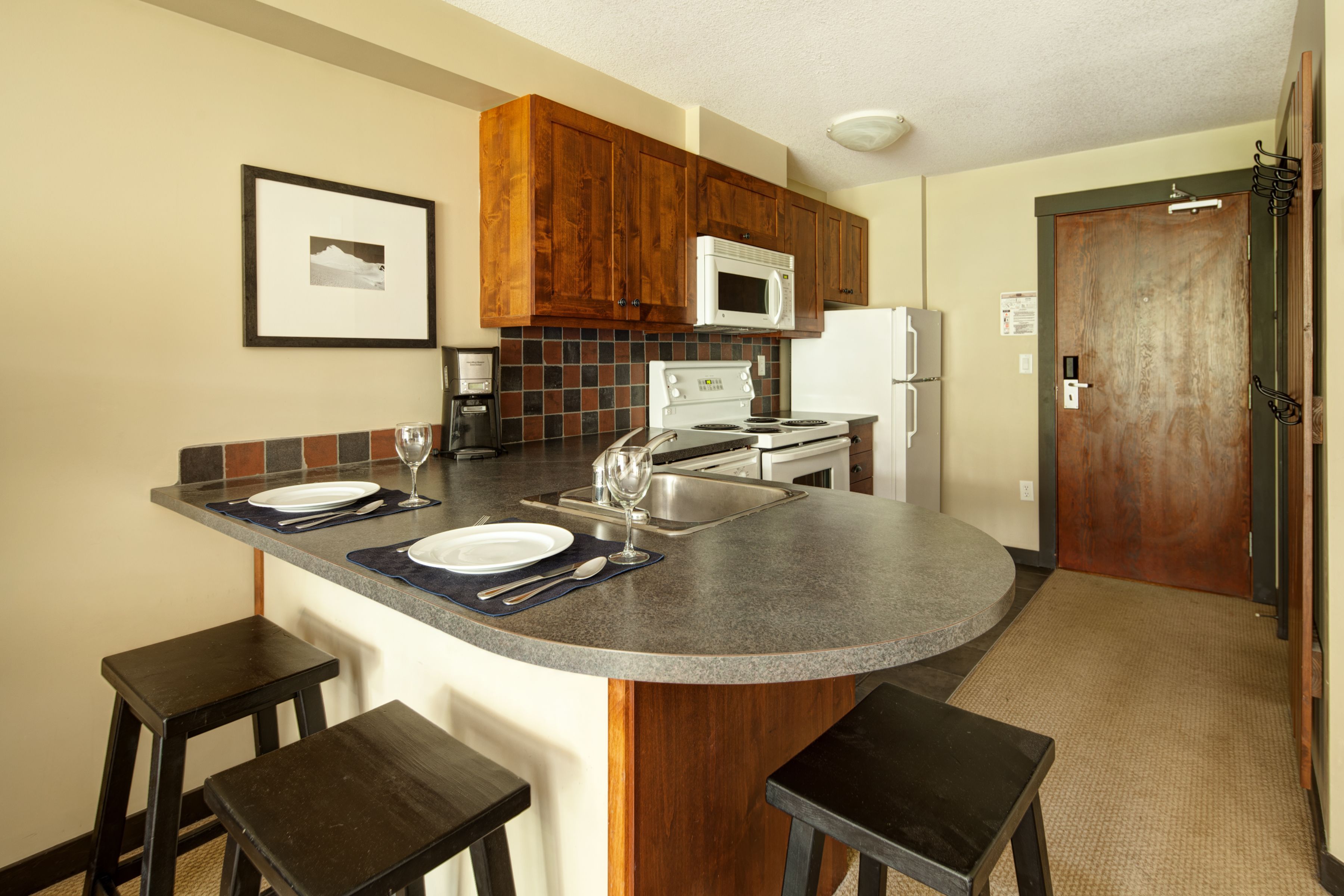 A kitchen with wooden cabinets, a microwave, a stove, and a refrigerator. There are bar stools and a small island with a sink.