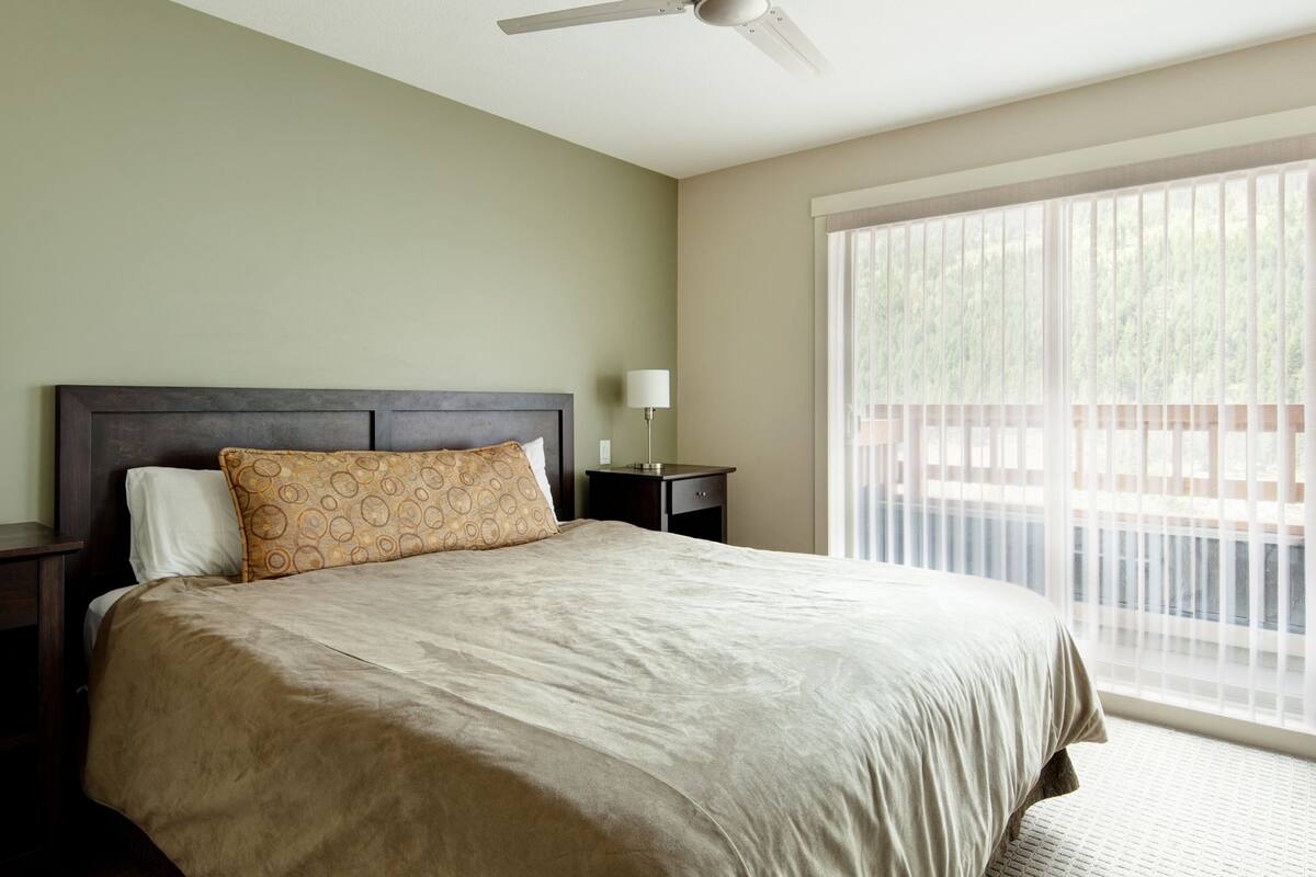A bedroom with a large bed, a nightstand, a lamp, and a window with blinds.