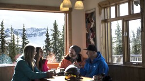 A group of people sitting at a wooden table inside a room with large windows overlooking a snowy mountain landscape. - Panorama Mountain Resort - Premium Condos and Townhomes (Panorama)